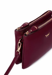 Burgundská kožená crossbody taška s dvěma zipovými přihrádky, zlatými kovovými prvky a odnímatelným popruhem. Hladká textura s logem.