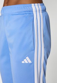 Ανοιχτό μπλε αθλητικά παντελόνια με λευκές ρίγες στα πλάγια, ελαστική μέση και μικρό λευκό λογότυπο. Λείος υφασμάτινος χαρακτήρας με χαλαρή σχεδίαση.