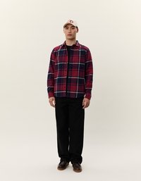 Rød og blå ternet flannel skjorte med lange ærmer, sorte bukser og brune sko. En beige kasket med rødt logo fuldender outfittet.