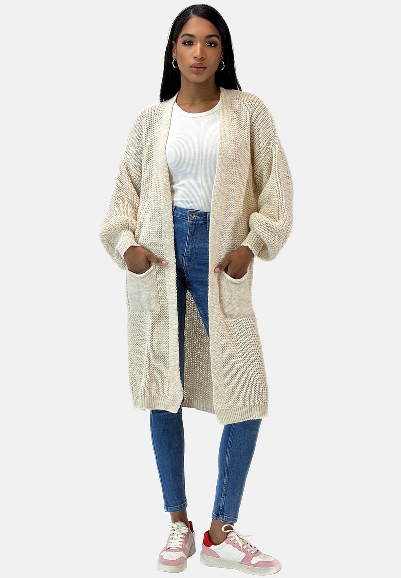 Lange beige cardigan van gebreide stof, met een relaxte pasvorm, open voorkant en twee voorzakken. Gecombineerd met blauwe jeans en sneakers.