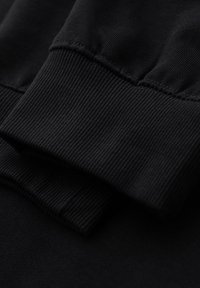 Svart sweatshirt tyg med ribbade ärmar och subtila sömnadsdetaljer samt en mjuk, texturerad yta. Närbild visar lager-på-lager-effekten.