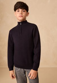 Tume sinine pusa kõrge krae ja ees oleva tõmblukuga, valmistatud pehmest kudumkangast. Sobitatud ruuduliste pükstega. Lihtne disain.