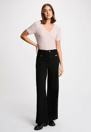 Donna con capelli castani alla lunghezza delle spalle indossa una maglietta rosa chiaro a maniche corte con scollo a V, pantaloni neri a gamba larga con tasche frontali e scarpe nere.