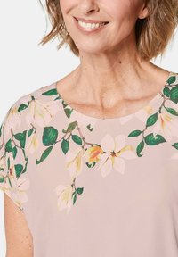 Bluse mit Blumenmuster, die einen hellrosa Hintergrund mit grünen Blättern und gelben Blumen zeigt. Rundhalsausschnitt und kurze Ärmel. Weiches Stoffgefühl.