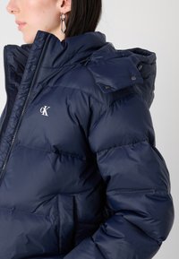 Marineblå pufferjakke med høy krage, glidelås foran, trykknapper og logo. Har kviltet tekstur og sidelommer.