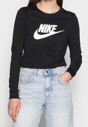 Czarna koszula z długim rękawem z białym logo Nike, zestawiona z jasnoniebieskimi dżinsami z wysokim stanem, ręka w kieszeni, stojąca na jednolitym tle.