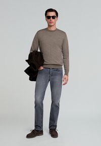 Brązowy sweter, szare dżinsy i brązowe buty; trzyma ciemny płaszcz. Okulary przeciwsłoneczne; prosty design o dopasowanym kroju. Gładka tekstura.
