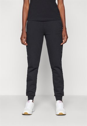 Osoba ubrana w czarne spodnie dresowe i białe trampki stojąca na tle jednolitego jasnego tła.