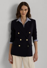 Marineblauwe dubbelgevoerde blazer met gouden knopen, geknipte revers en opgestikte zakken. Daaronder is een blauw-wit gestreept overhemd met kraag zichtbaar.