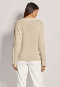 Beiger Strickpullover mit herabgesetzten Schultern, geripptem Saum und Ärmelverzierungen. Weicher Textur mit lockerer Passform, getragen mit weißen Hosen.