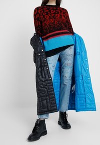 Czerwony i czarny sweter w panterkę z niebieskim pasem, zestawiony z jasnymi, przetartymi dżinsami, czarną pikowaną kurtką i czarnymi butami.