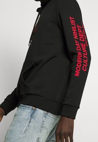 Sudadera negra de algodón con texto rojo a lo largo de la manga derecha. Bolsillo canguro en la parte frontal y puños acanalados en el dobladillo.