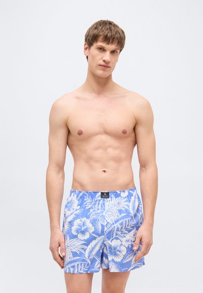 Νεαρός άνδρας με κοντά καστανά μαλλιά που φοράει μπλε και λευκά μαγιό με λουλουδάτο μοτίβο, στέκεται μπροστά σε απλό φωτεινό φόντο.