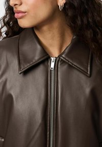 Veste en cuir synthétique marron avec un col pointu, dotée d'une fermeture éclair métallique et d'une texture lisse, mettant en valeur des détails de couture.