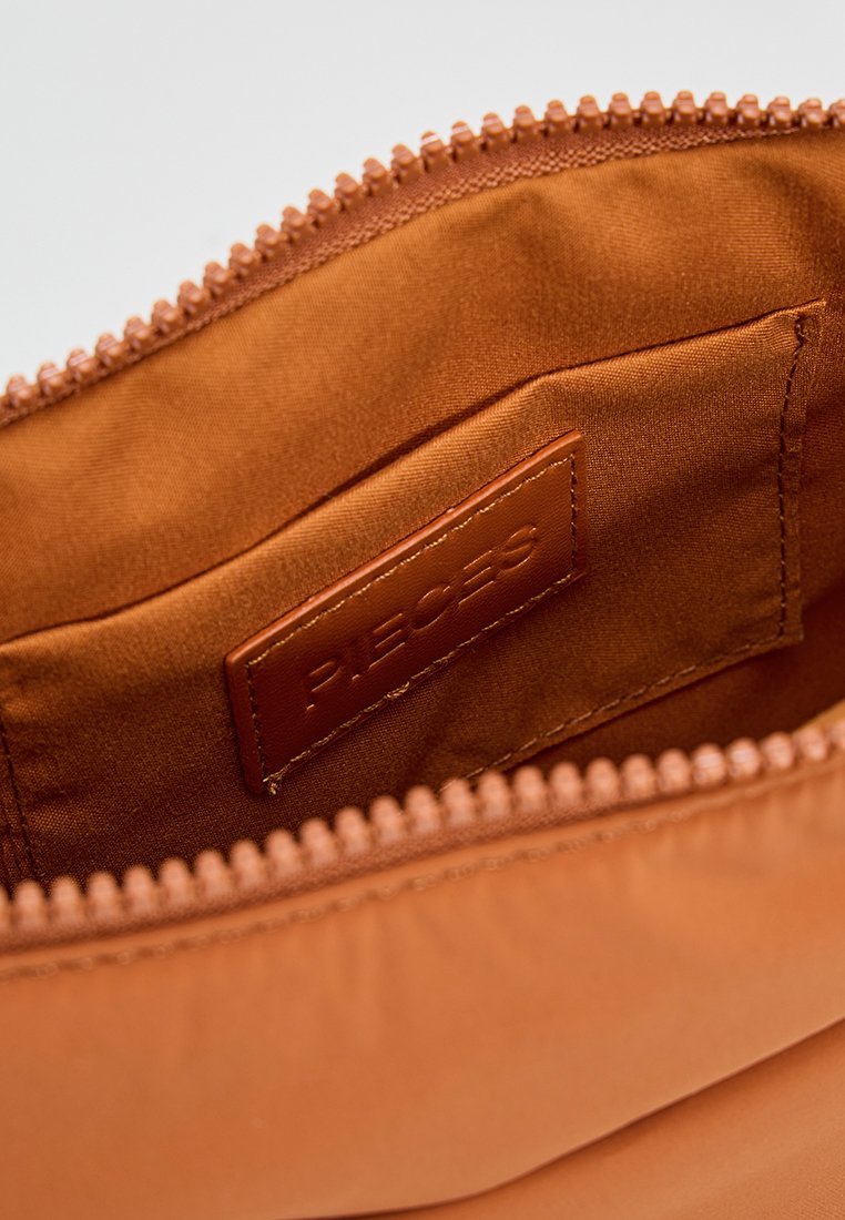 Pochette marron zippée avec une texture lisse, dotée d'une étiquette en cuir à l'intérieur sur laquelle est inscrit "P.I.E.C.E.S." L'intérieur est doublé d'un tissu doux.