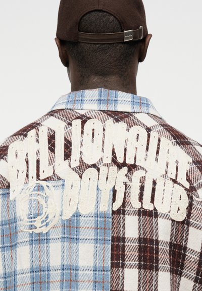 Muž nosící hnědou čepici a kostkovanou košili s nápisem "Billionaire Boys Club" bílými písmeny na zádech.