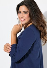 Marinblå långärmad tröja med rund halsringning och mjuk, slät textur. Visad på en person med mörkt, vågigt hår som ler milt.