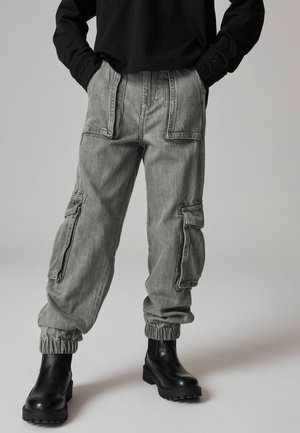 Pantalons cargo gris avec poches latérales, poignets élastiques et coupe décontractée, associés à des bottines noires à la cheville. Design simple avec un tissu légèrement texturé.