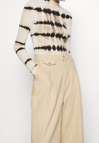 Top de manga larga en beige con estampado tie-dye y rayas horizontales negras, combinado con pantalones de tiro alto en beige claro con bolsillos delanteros.