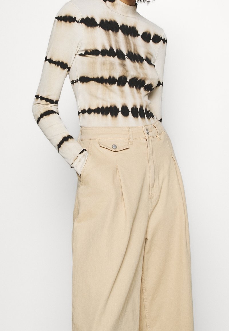 Top de manga larga en beige con estampado tie-dye y rayas horizontales negras, combinado con pantalones de tiro alto en beige claro con bolsillos delanteros.