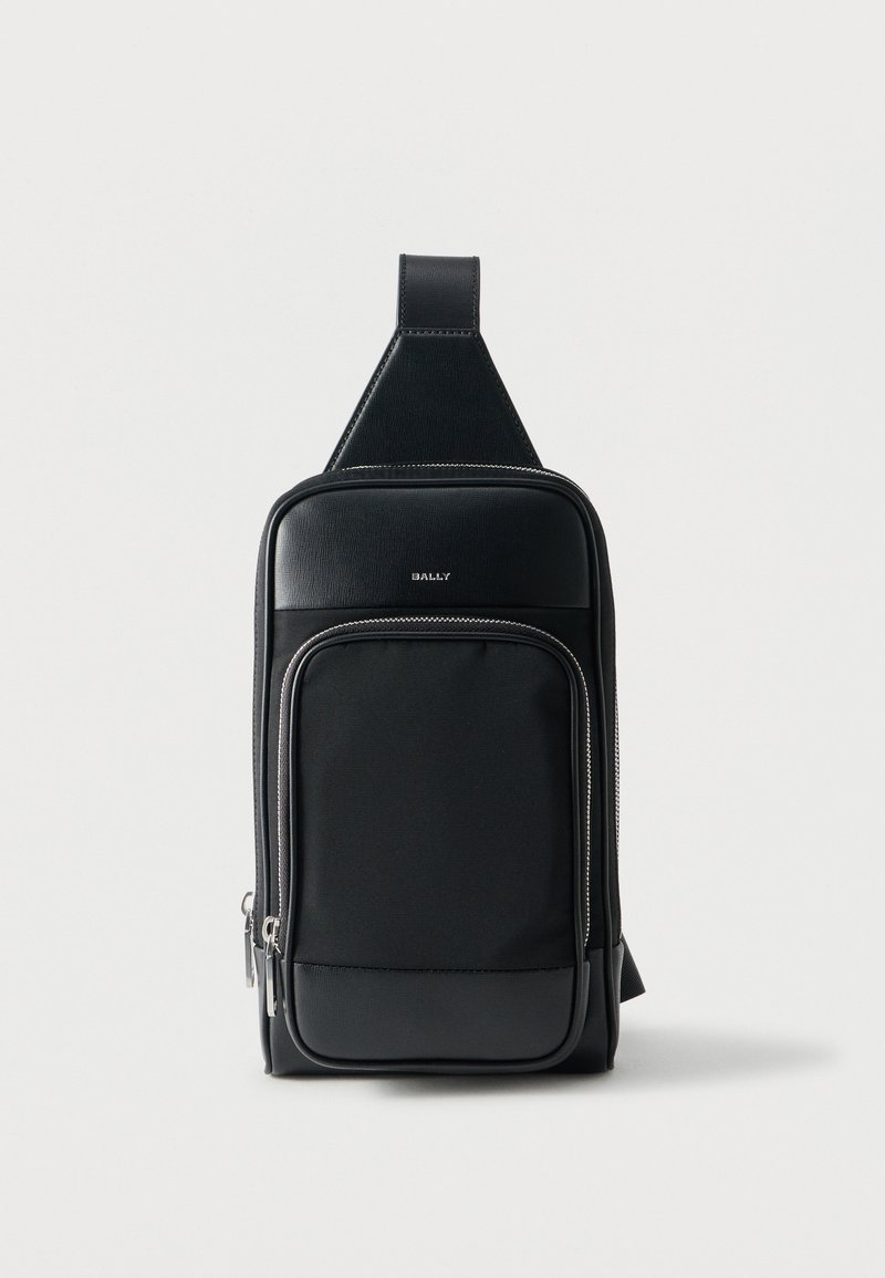 Sac à dos noir à bandoulière avec deux compartiments zippés et une sangle rembourrée, arborant un petit logo métallique "BALLY" à l'avant.