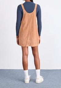 Braunes Corduroy-Latzkleid mit breiten Trägern, einem Knopfdetail auf der Rückseite, getragen über einem langärmeligen marineblauen Oberteil und weißen Sneakers.