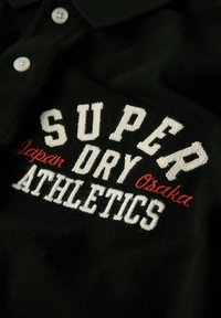 Maglietta polo nera con testo ricamato bianco e rosso: "SUPER DRY JAPAN OSAKA ATHLETICS." Completata con bottoni bianchi.