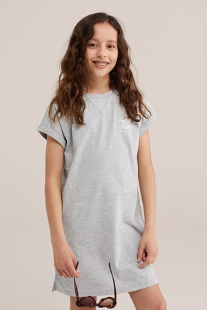 Chica con pelo largo y rizado que lleva un vestido gris de manga corta, sosteniendo gafas de sol con montura carey, sonriendo con brackets sobre un fondo beige.