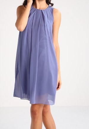 Femme portant une robe sans manches, longueur genou, en chiffon violet clair avec des plis au niveau du décolleté, debout devant un fond uni.