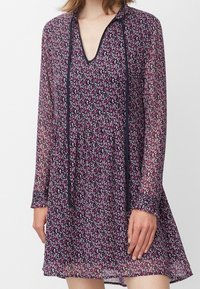 Lila blomklänning med långa ärmar, djup V-ringning och kontrasterande marinblå band. Har en lätt, genomskinlig tyg och inringad midja.