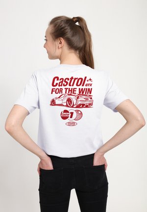 Jeune femme avec queue de cheval brune portant un t-shirt blanc avec l'inscription rouge "Castrol GTX FOR THE WIN" et un graphique de voiture de course au dos, pantalon noir.
