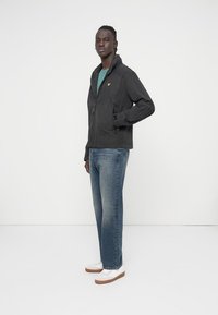 Black zip-up jacket with a textured finish, single pocket, worn over a teal shirt, paired with blue jeans and white sneakers.