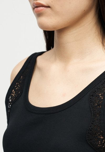 Γυναίκα που φοράει μαύρο ριμπαντό τοπ με δαντελένιες λεπτομέρειες στους ώμους, εμφανίζοντας το λαιμό και το επάνω μέρος του στήθους της.