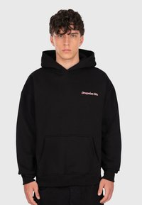 Young man with curly hair wearing a black hoodie with "Dropsize Clo." text in pink on the chest, standing against a plain background.