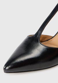 Scarpa nera in pelle con tacco alto, dalla punta appuntita e lati a taglio. La texture liscia e la fodera interna color cammello sono visibili.