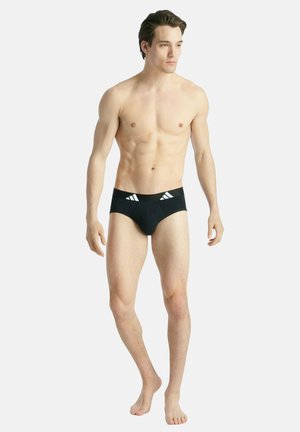 Schwarze Schwimmbriefs mit enger Passform, versehen mit weißen diagonalen Streifen am Bund. Glattes Material, minimalistisches Design, geeignet zum Schwimmen.