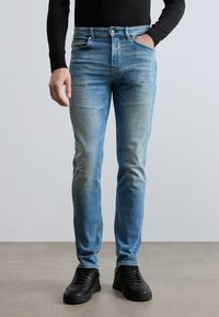 Svetlomodré riflové nohavice s úzkym strihom, jemným blednutím a opotrebovaným detailom na ľavom kolene, kombinované s čiernymi teniskami.