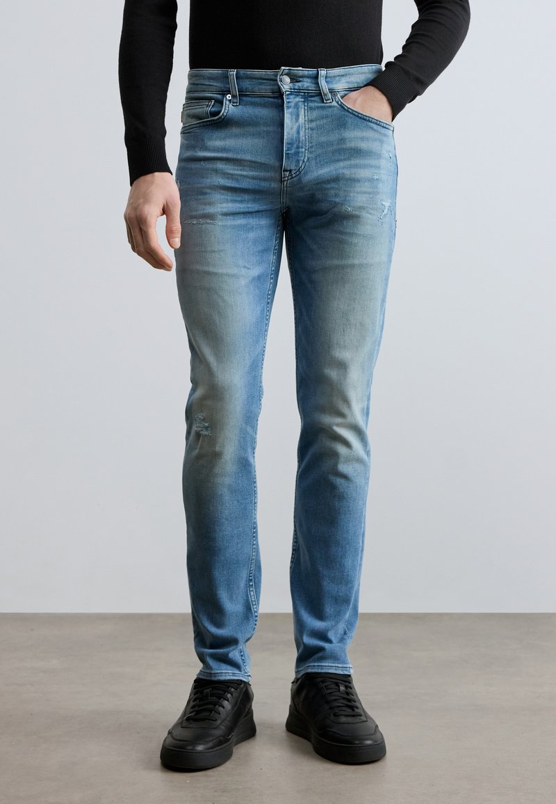 Svetlomodré riflové nohavice s úzkym strihom, jemným blednutím a opotrebovaným detailom na ľavom kolene, kombinované s čiernymi teniskami.