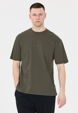 Mand med kort hår iført en olivengrøn oversized t-shirt og sorte bukser, stående mod en hvid baggrund.