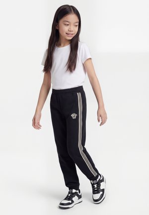 Noor tüdruk kannab valget t-särki, musti trennipükse beežide triipudega ja must-valgeid spordijalatsid, seisab valgel taustal.