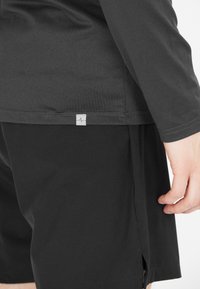 Mustapaita, joka on valmistettu pehmeästä materiaalista, pitkät hihat ja sivuaukot, yhdistetty mustiin lyhyisiin housuihin. Paidassa on pieni logotagi.