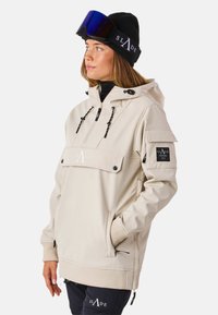 Beżowa kurtka z kapturem, z kieszenią z przodu, sznurkiem i czarnymi akcentami, w połączeniu z czarną czapką beanie i dużymi goglami narciarskimi.