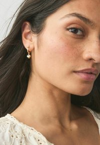 Orecchini a cerchio in oro con un piccolo ciondolo a forma di cuore, caratterizzati da una superficie lucida e un design semplice, indossati da una modella con pelle chiara.