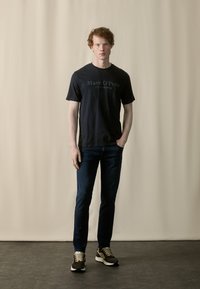 Navy cotton t-shirt with logo, paired with dark denim jeans and multicolored sneakers. The background is a soft beige.