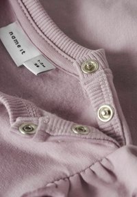 Lichtroze katoenen kledingstuk met een ribbelkraag, drukknoopsluiting en een gestikte ruches aan de zoom. Het label zegt "name it," maat 56.