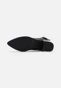 Zapato de tacón alto de ante negro con puntera afilada, que cuenta con una suela texturizada y una correa ajustable con un acento de metal plateado.