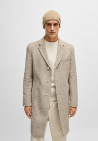 Cappotto beige a trama con rever e tre bottoni. Indossato sopra un maglione chiaro, abbinato a un berretto beige a coste.