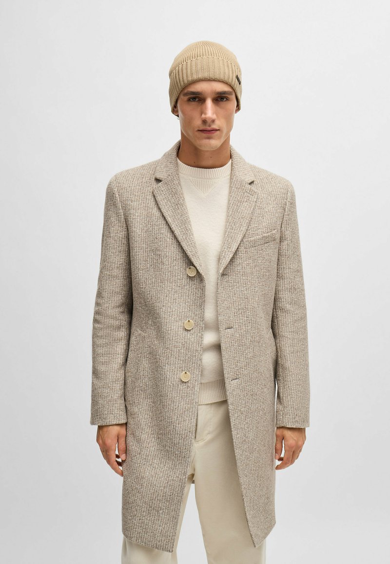 Cappotto beige a trama con rever e tre bottoni. Indossato sopra un maglione chiaro, abbinato a un berretto beige a coste.