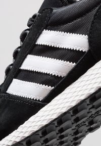 Lähikuva mustasta tennarista, jossa on valkoiset vaaleahampaiset raidat ja teksturoitu valkoinen välipohja mustalla urakumipohjalla.