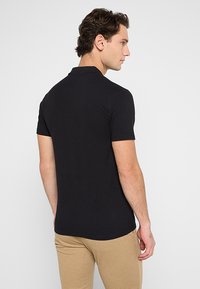 Czarna koszulka polo z krótkim rękawem, z kołnierzykiem i gładką teksturą. Połączona z jasnobeżowymi spodniami, noszonymi z tyłu.